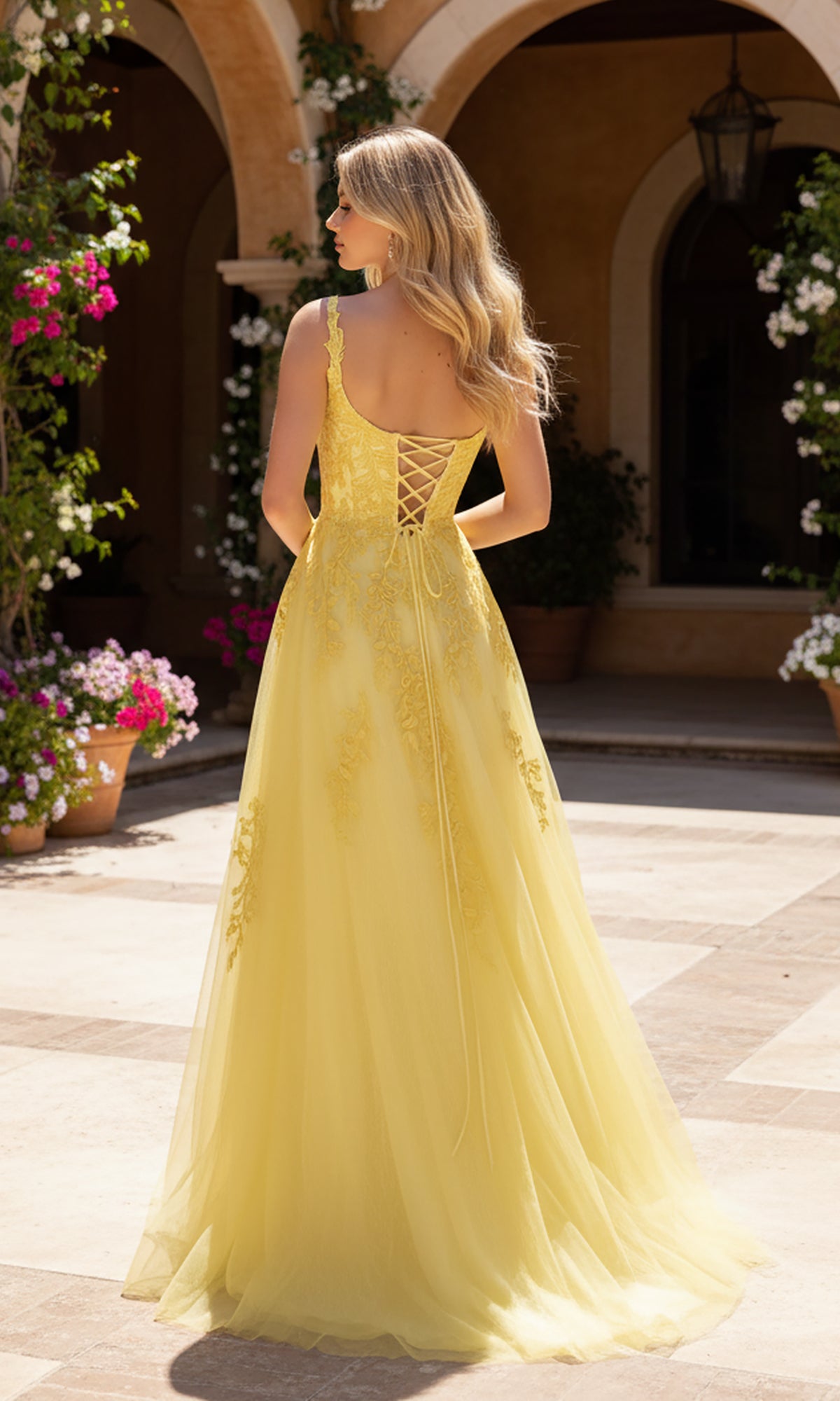 A woman with long blonde hair models the PromGirl Exclusive 250233, a yellow a-line prom dress with lace appliques and a lace-up back, outdoors on a stone patio surrounded by potted flowers and arches.